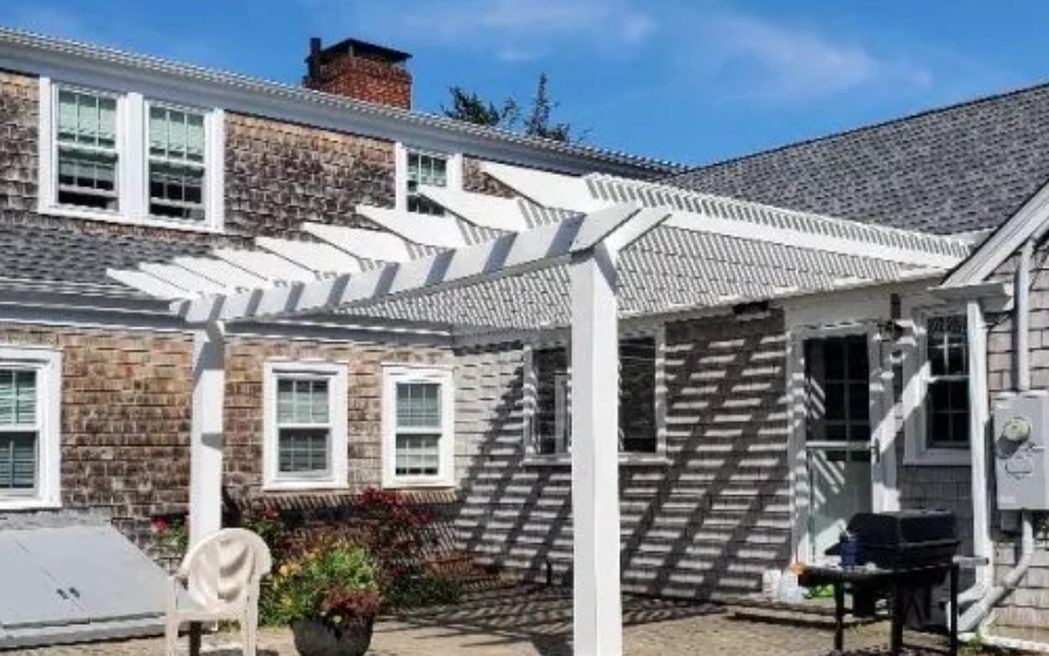 A white pergola tied into a shingled house above a paver patio